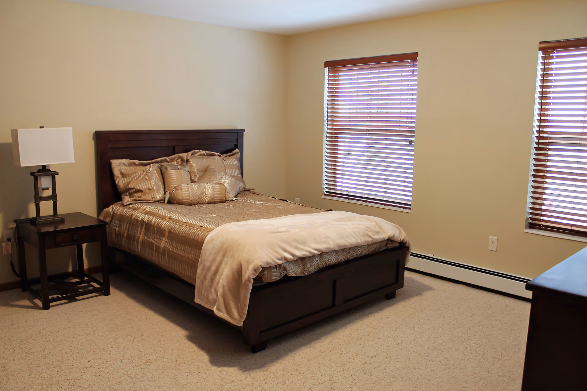 A bedroom with a neatly made bed featuring a dark wooden frame and beige bedding. There are two windows with wooden blinds on the right wall, a dark wooden nightstand with a lamp on the left side of the bed, and a beige carpeted floor.