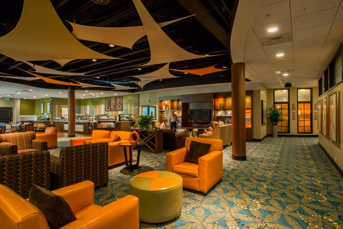 A spacious and well-lit lounge area with comfortable orange and patterned armchairs, a round ottoman, and a carpet with a leaf pattern. The ceiling features decorative fabric panels in shades of beige, orange, and blue. In the background, there is a café counter labeled 'Vitality' with menu boards and display cases. To the right, a hallway leads to doors marked 'Day Spa'.
