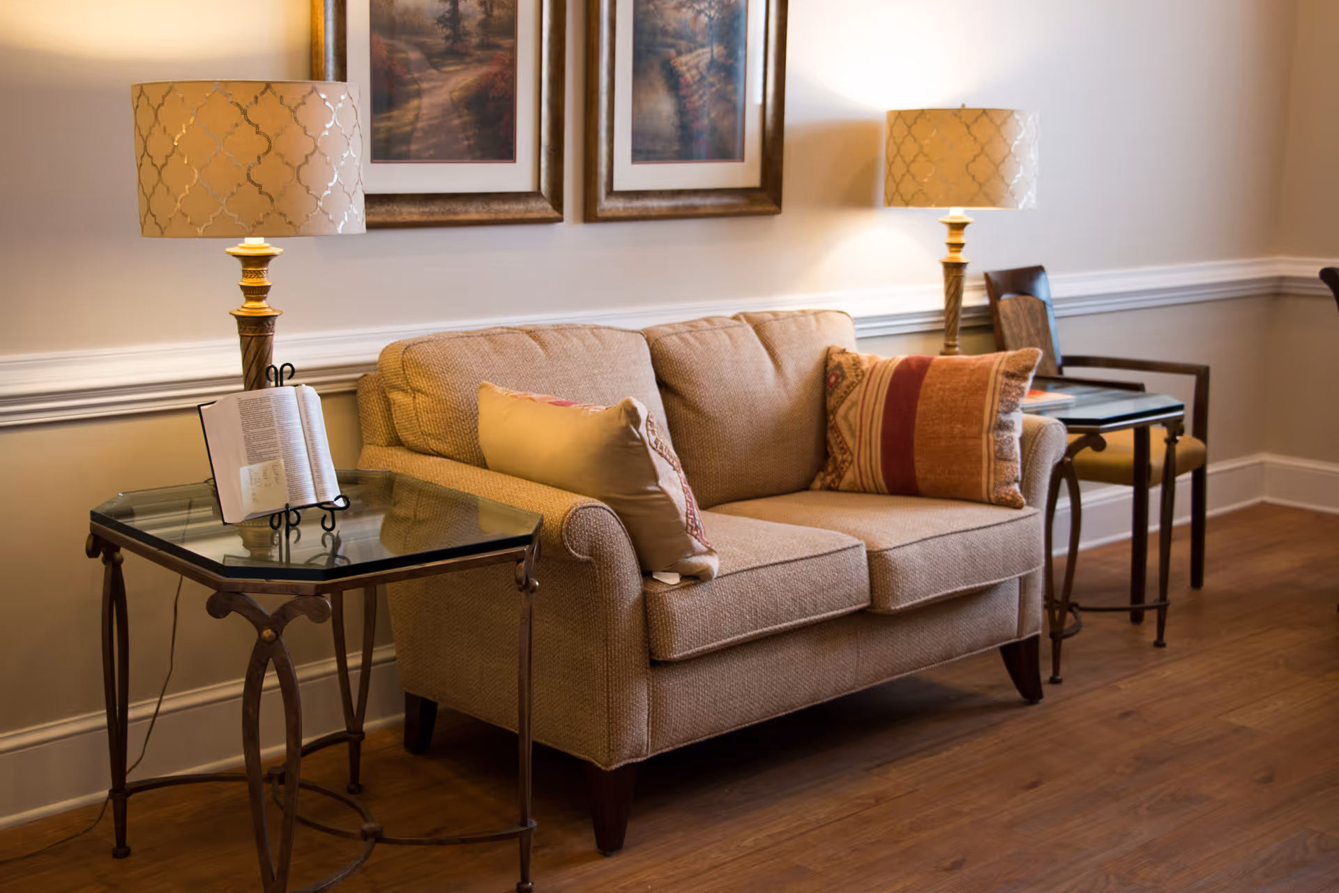 A cozy living room area featuring a beige loveseat with two decorative pillows, flanked by two glass-top side tables each holding a lamp with a patterned shade. Two framed landscape paintings hang on the wall above the loveseat. The room has wooden flooring and neutral-colored walls with white trim.