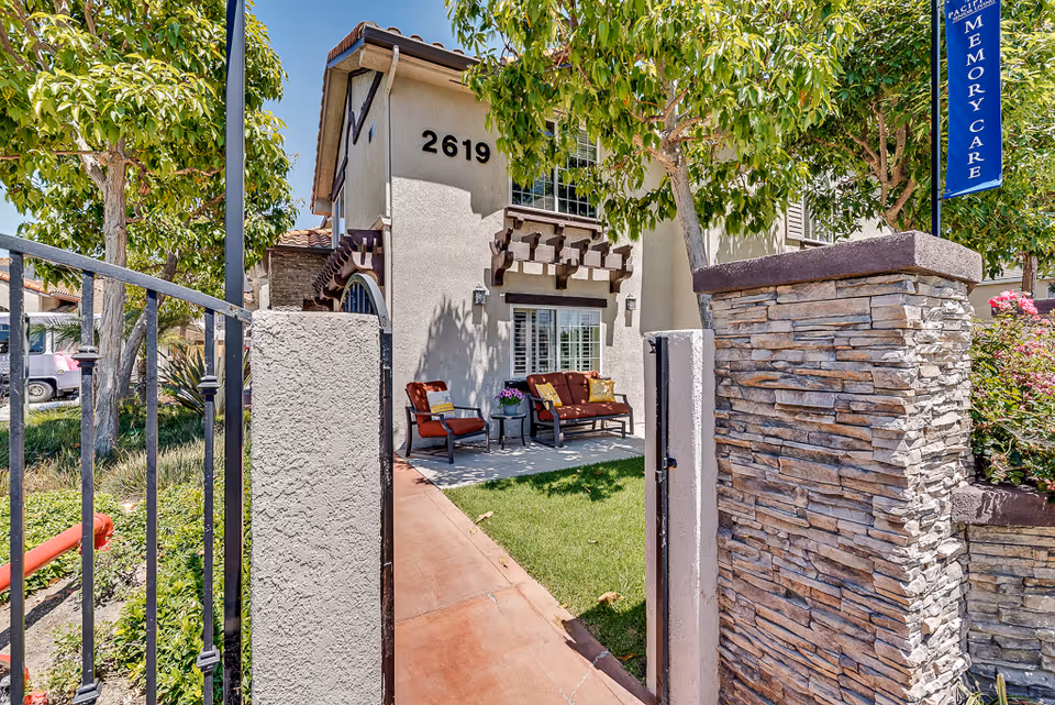 Gated front entrance and small patio seating area of a senior living building numbered 2619 with trees and a stone pillar.