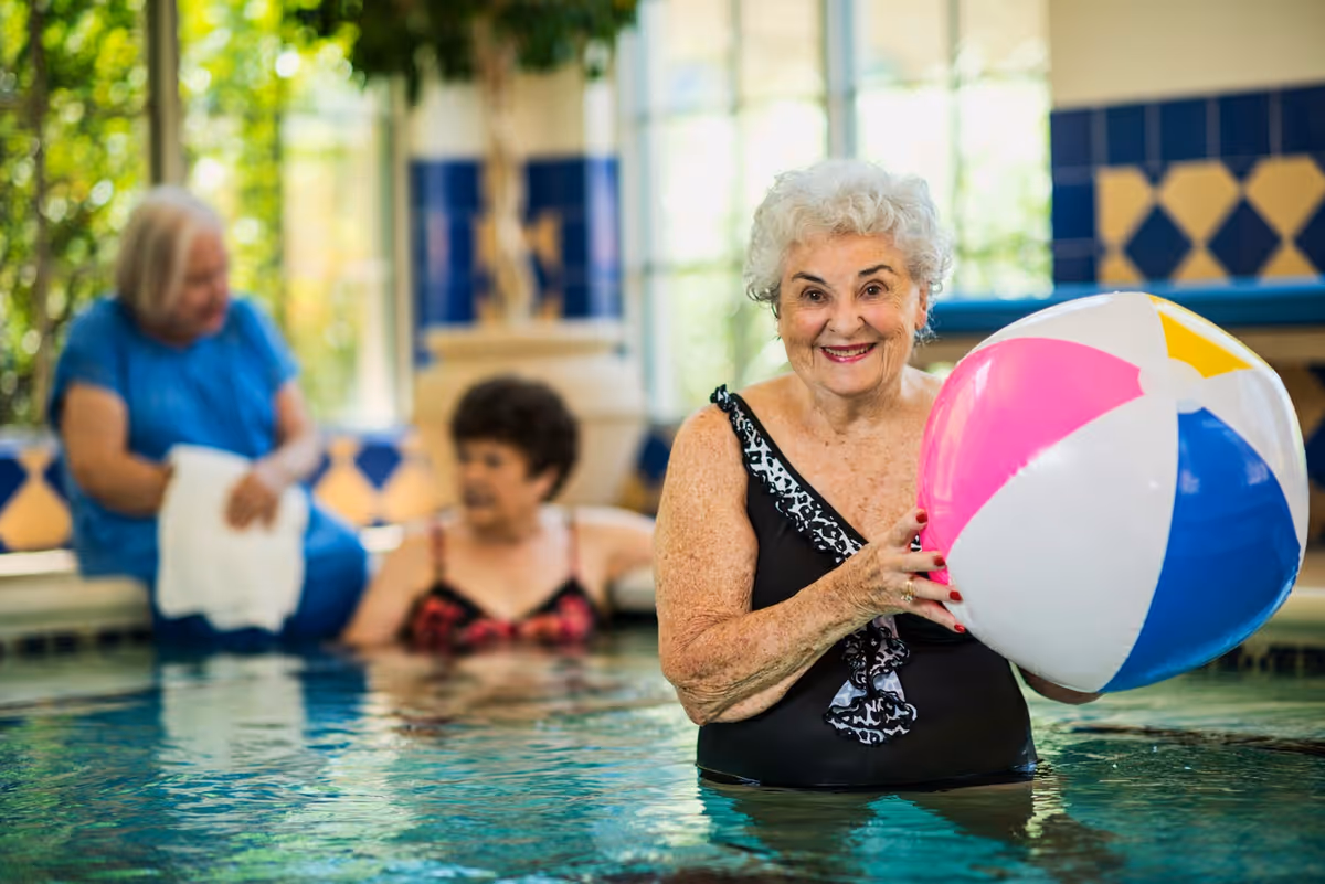 An elderly woman in a black swimsuit holding a colorful beach ball while standing in an indoor pool. Two other elderly women are in the background, one sitting on the edge of the pool with a towel and the other partially submerged in the water. The pool area has blue and yellow tiled walls and large windows letting in natural light.