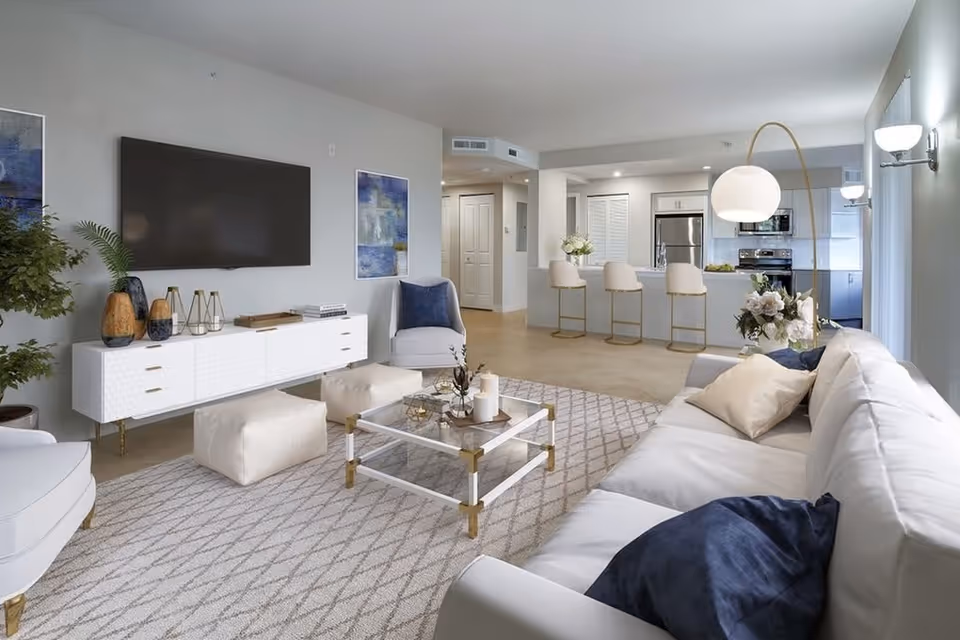 A modern living room with a large white sofa adorned with blue and beige pillows, a glass coffee table with decorative items, two white poufs, and a white media console with vases and books. A flat-screen TV is mounted on the wall above the console. The room opens into a kitchen area with a breakfast bar and three beige bar stools. The space is well-lit with natural light from large windows and a floor lamp with a round white shade.
