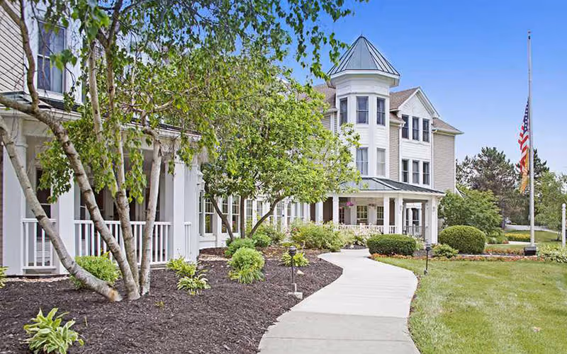Exterior view of a senior living facility with a curved concrete walkway leading to the entrance. The building has white siding, multiple windows, and a turret-like architectural feature. There are trees, shrubs, and well-maintained landscaping along the walkway, and an American flag is flying at half-mast on a flagpole in the grassy area to the right.