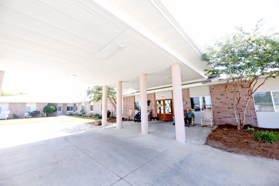 Covered entrance area of a senior living facility with brick walls, several pillars supporting the roof, rocking chairs, a wheelchair, and two people sitting near the entrance door. There are trees and landscaping around the building.