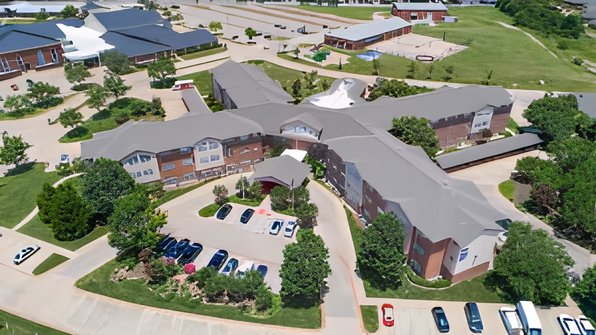 Aerial view of Morada Cedar Hill senior living facility showing a large building with a gray roof surrounded by green lawns, trees, and parking lots with several cars parked.