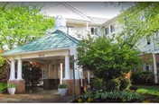 Entrance to a senior living facility with a covered driveway supported by white columns and a green roof. The building is light-colored with multiple windows and surrounded by green trees and landscaped bushes.