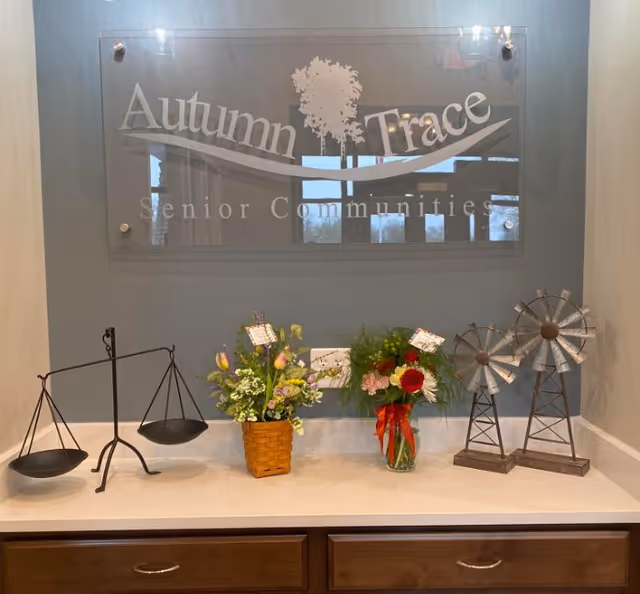 Reception display with an 'Autumn Trace Senior Communities' wall sign above a counter holding two floral arrangements, decorative balance scales, and metal windmill sculptures.