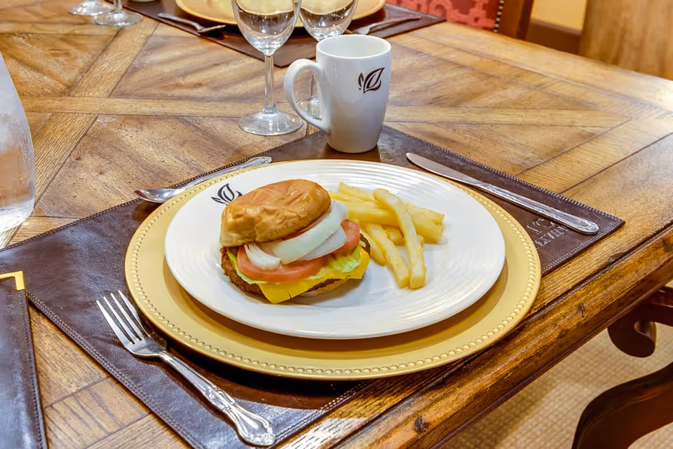 A wooden dining table set with a plate containing a cheeseburger with lettuce, tomato, and onion, accompanied by French fries. The plate is placed on a gold charger plate and a brown leather placemat. There is a white mug with a leaf logo, a fork, knife, and two empty wine glasses on the table.