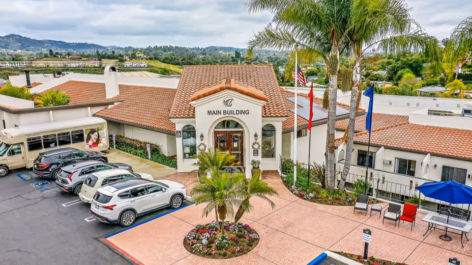 Exterior view of the main building at Rancho Vista Senior Living, featuring a tiled roof, an entrance with an archway, several parked cars, a shuttle bus, palm trees, flags, and outdoor seating with a blue umbrella.