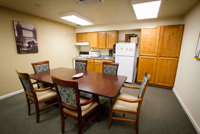 A small kitchen and dining area with a wooden table surrounded by six chairs with patterned upholstery. The kitchen features wooden cabinets, a white refrigerator, a microwave, and a stove. The room has beige walls and a carpeted floor.