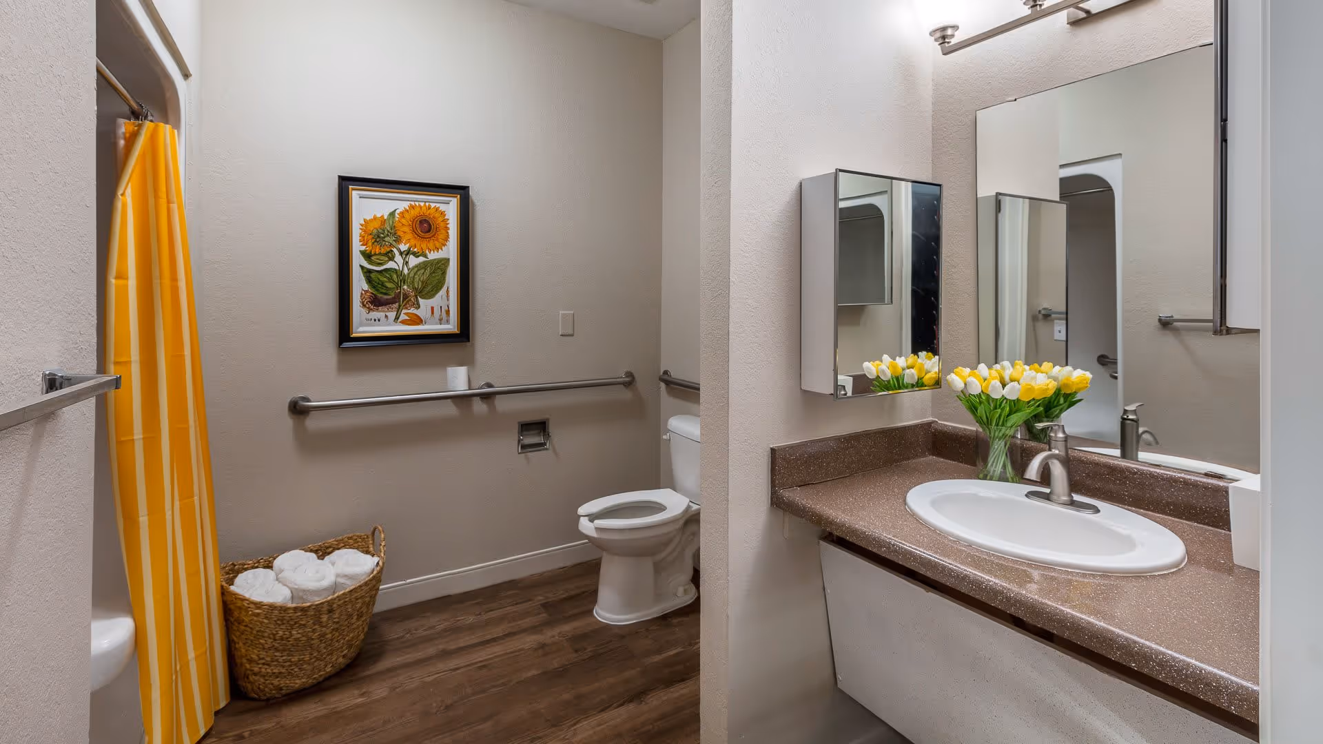 A clean and accessible bathroom with a white toilet, a brown countertop with a white sink, a mirror above the sink, a vase of yellow and white tulips on the counter, a yellow striped shower curtain, a basket with rolled white towels, a framed sunflower picture on the wall, and grab bars installed near the toilet and shower area.