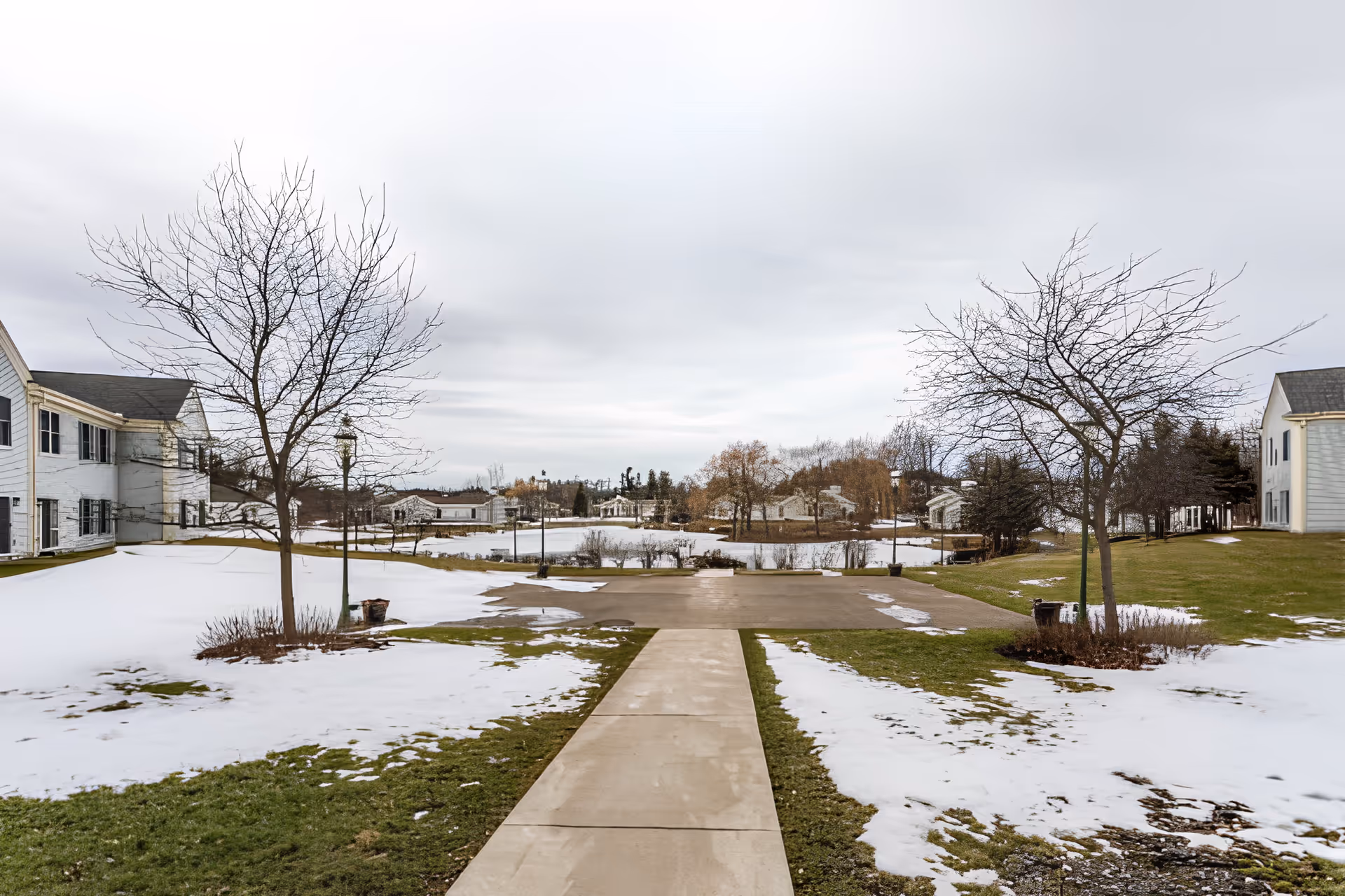 A winter outdoor scene at Elison Independent Living of Orchard Glen showing a concrete walkway leading through a partially snow-covered grassy area with leafless trees on either side. Residential buildings are visible on the left and right sides, and more buildings and trees are seen in the background under a cloudy sky.