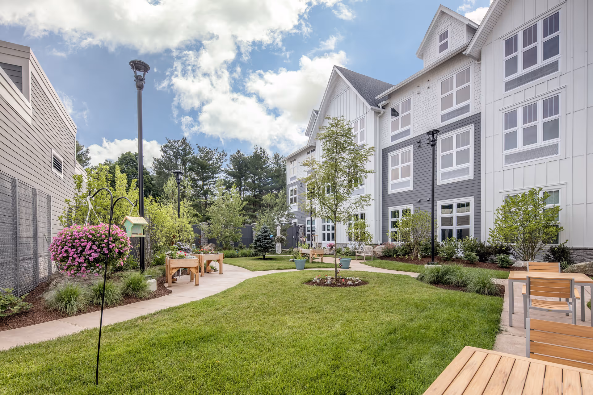 Sunny outdoor courtyard with green lawn, trees, and seating beside a white multi-story building