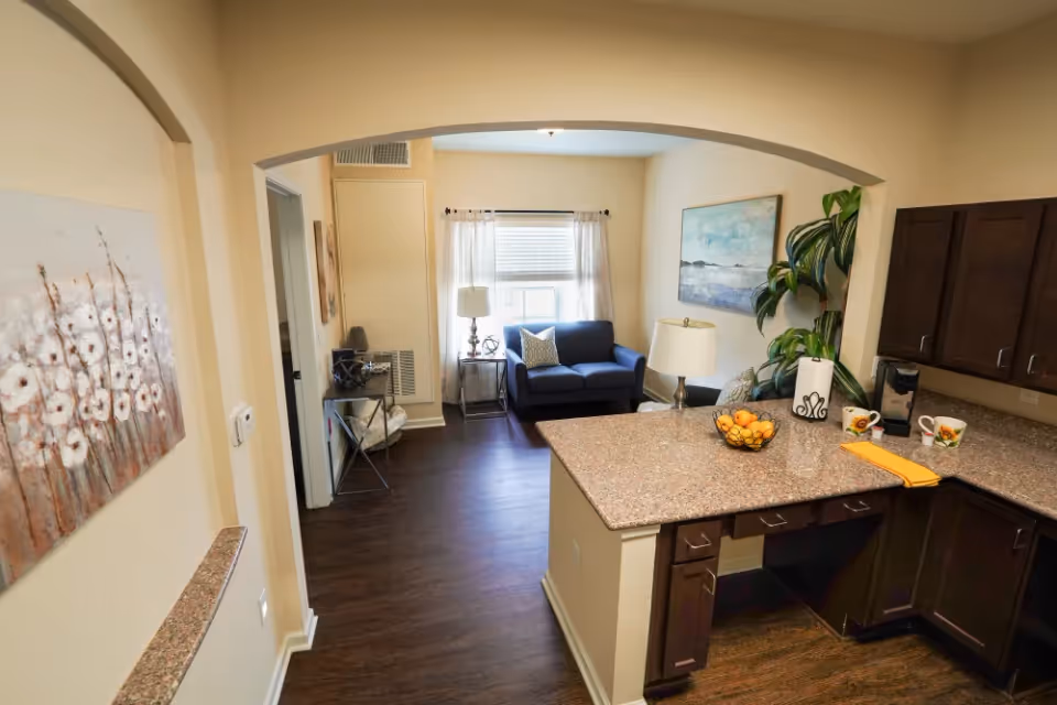Interior view of a senior living facility showing a kitchen area with granite countertops, dark wood cabinets, a coffee maker, and a bowl of oranges. Adjacent to the kitchen is a living room with a blue loveseat, side tables with lamps, a large plant, and wall art. The space has wooden flooring and light-colored walls.