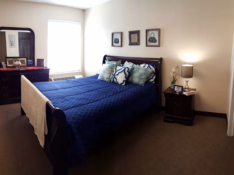 A bedroom with a dark wooden bed frame and a blue quilted bedspread. The bed has several decorative pillows in shades of blue and white. On the right side of the bed is a dark wooden nightstand with a lamp, a small plant, a framed photo, and a book. On the left side of the room is a dark wooden dresser with a mirror, a red runner, and framed pictures. The walls are light-colored, and there are three framed pictures hanging above the bed. A window with blinds is on the left wall.