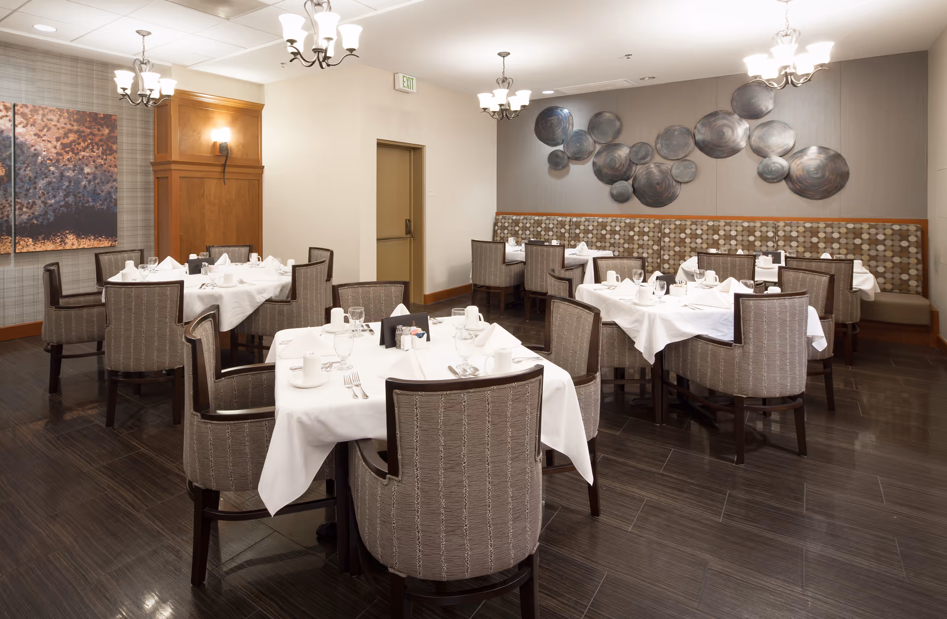 A dining room with several tables covered in white tablecloths, each set with cups, glasses, and silverware. The room features upholstered chairs, modern chandeliers, decorative wall art, and a patterned bench along one wall.