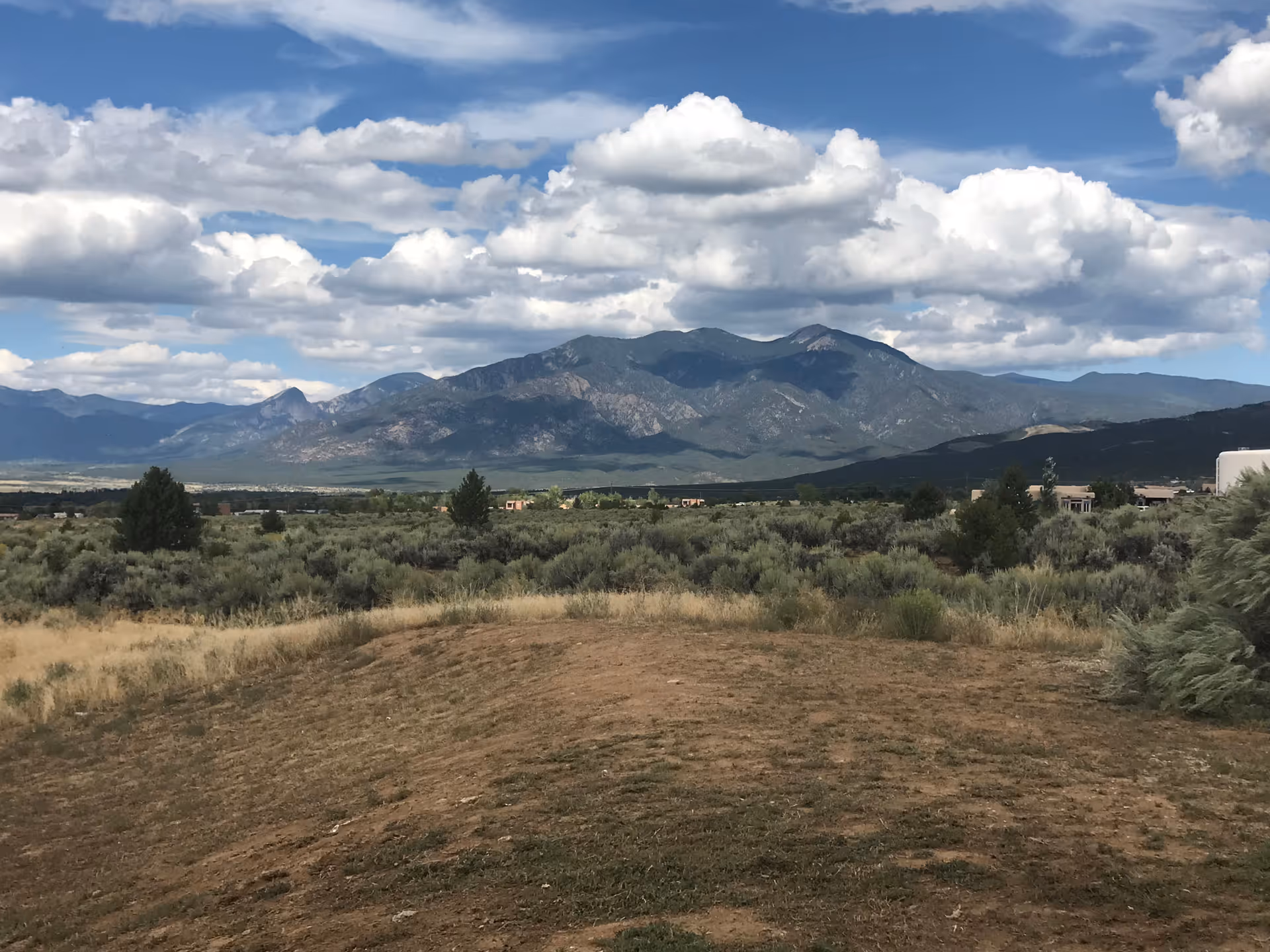 A scenic outdoor view of a mountainous landscape under a partly cloudy sky with shrubs and dry grass in the foreground.