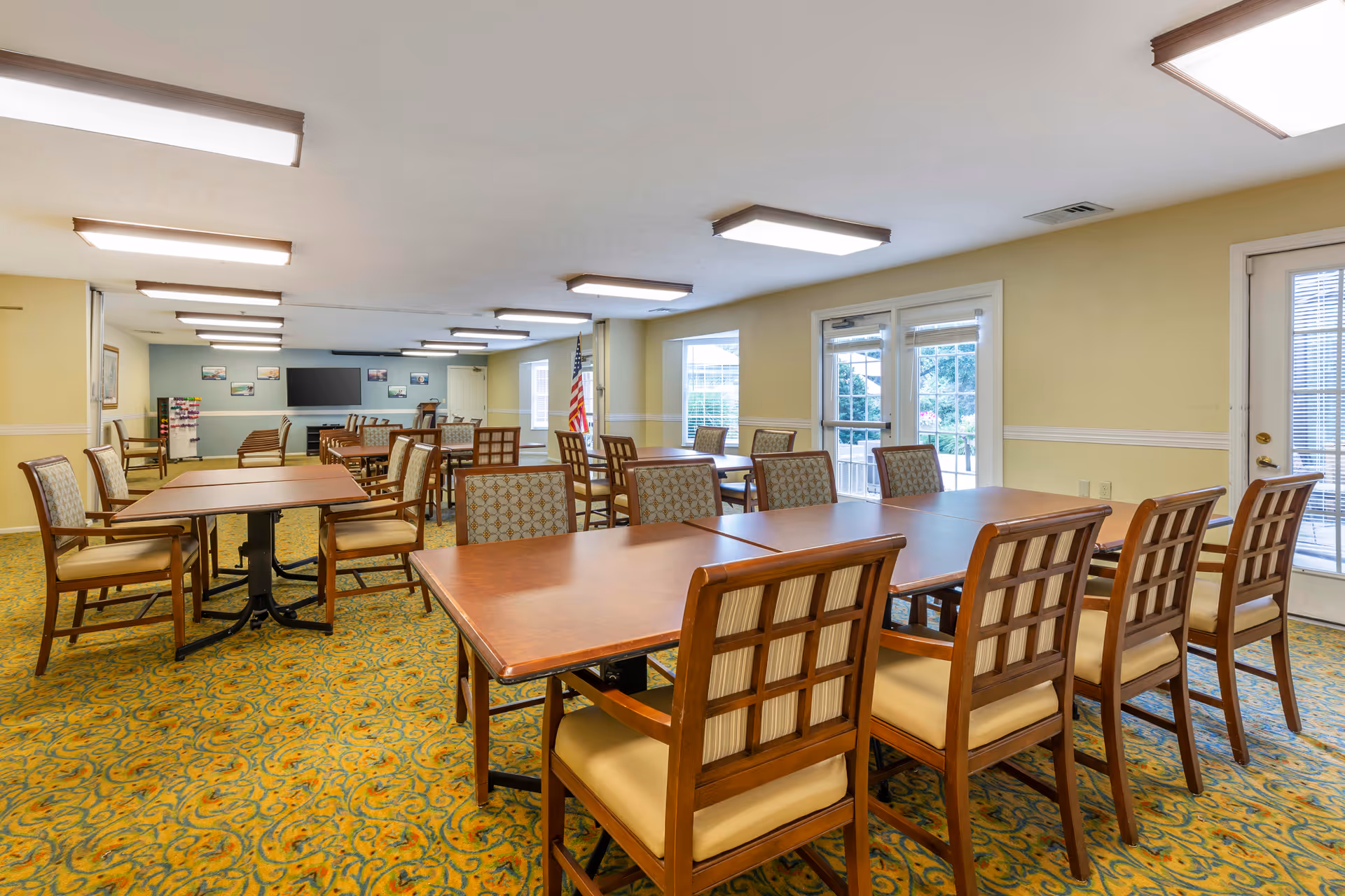 A spacious dining room with multiple wooden tables and cushioned chairs arranged neatly. The room has yellow walls, patterned carpet flooring, large windows, and glass doors letting in natural light. An American flag is visible near the back wall, which also features a large flat-screen TV and several framed pictures.