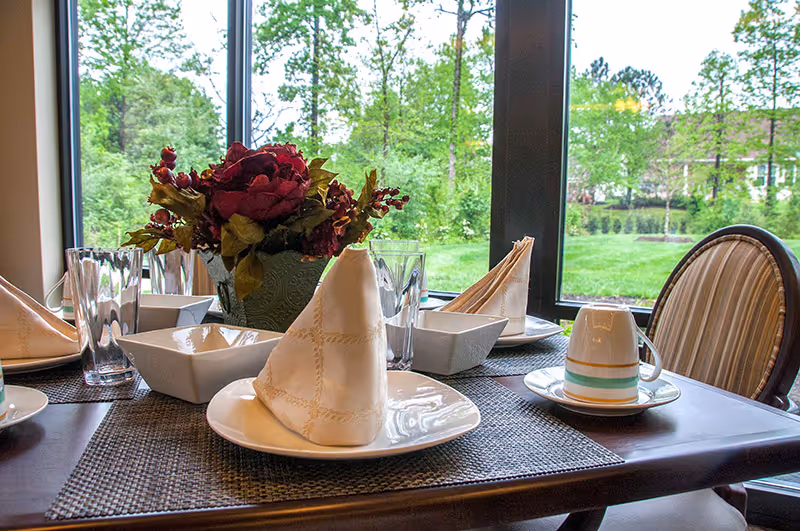 A dining table set with folded napkins, glasses, dishes and a floral centerpiece beside a large window overlooking green landscaping.