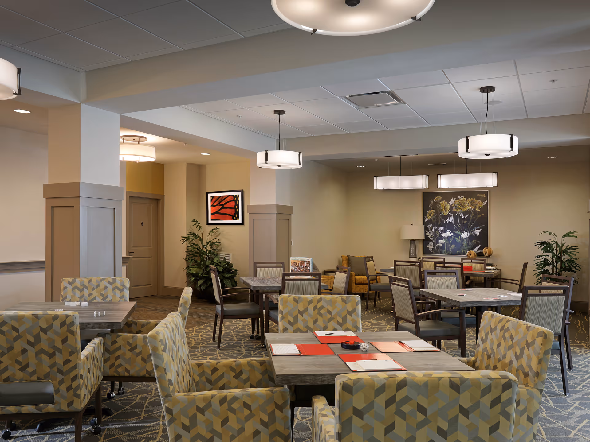 A well-lit communal dining and seating area with multiple tables, patterned armchairs, pendant lights, artwork, and potted plants.