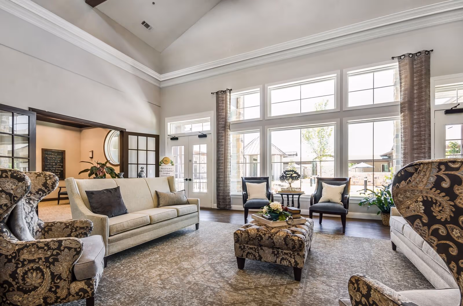 A bright and spacious living room with high vaulted ceilings and large windows letting in natural light. The room features a beige sofa with two pillows, two patterned armchairs, two dark wooden chairs with light cushions, and a patterned ottoman with books and a flower arrangement on top. There are sheer curtains on the windows and a door leading outside. The room has a neutral color palette with light walls and a patterned carpet.