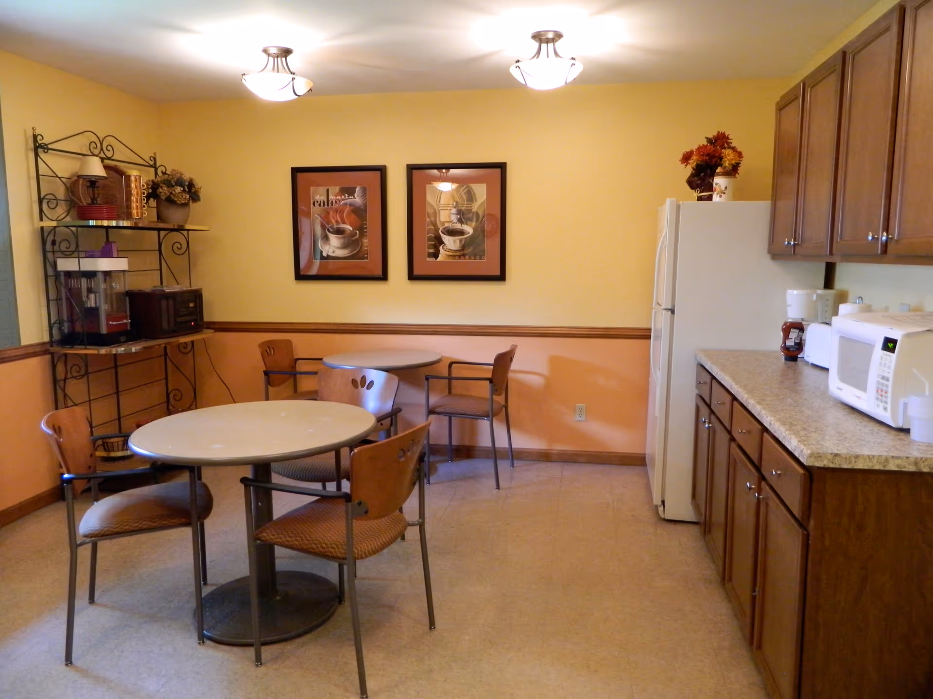 A small dining area with two round tables and several chairs. The walls are painted yellow and peach with a wooden chair rail. On the right side, there is a kitchen counter with a microwave, toaster, and refrigerator. On the left side, there is a metal shelving unit with a popcorn machine, microwave, and decorative items. Two framed pictures of coffee cups hang on the wall.