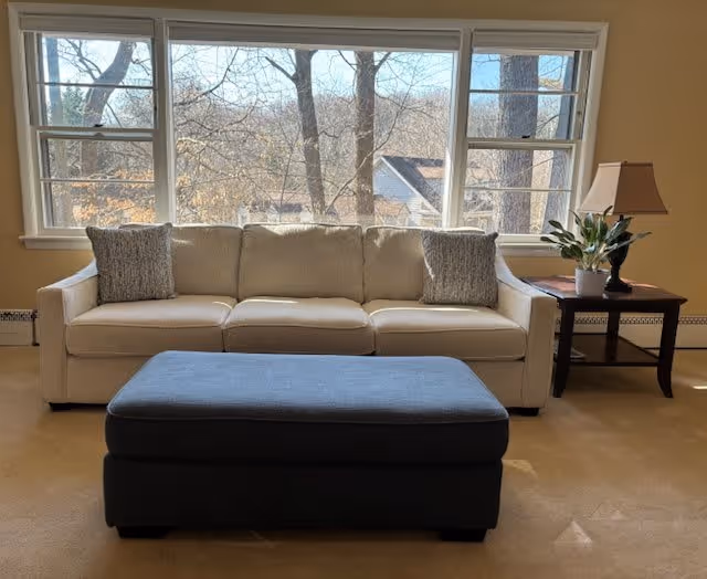 A beige sofa with two gray pillows is placed in front of a large window showing trees and houses outside. In front of the sofa, there is a dark gray ottoman. To the right of the sofa, there is a wooden side table with a lamp and a potted plant.