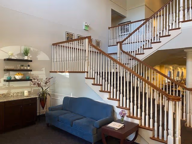 Interior view of a senior living facility featuring a blue upholstered sofa next to a wooden side table with a small flower vase and a book. Behind the sofa is a staircase with white spindles and wooden handrails leading to an upper floor. To the left, there is a dark wooden cabinet with granite countertop and three floating shelves decorated with plants, bowls, and decorative items. The walls are light-colored and there are framed pictures hanging near the staircase.