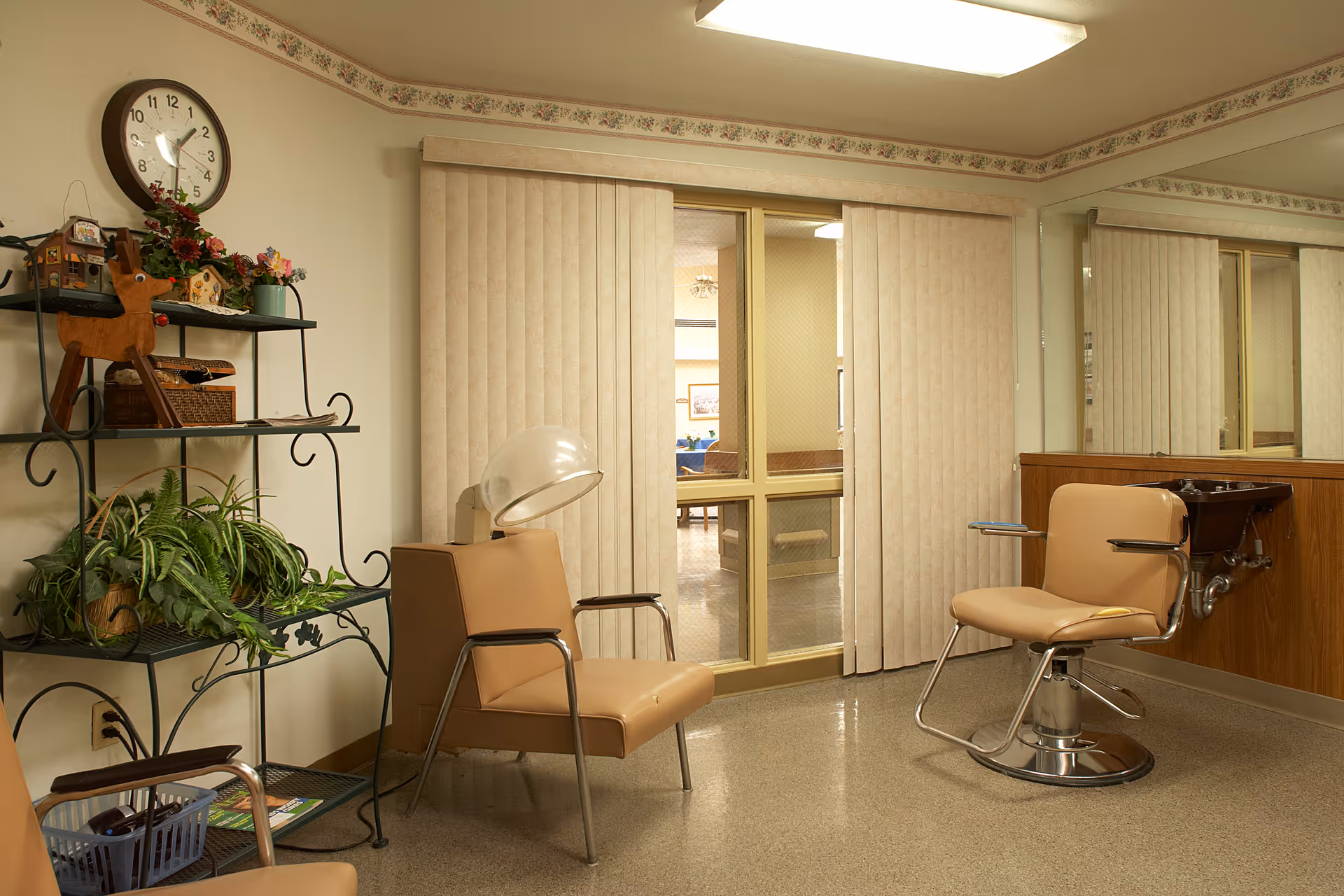 Interior of a salon area in a senior living facility with beige salon chairs, a hair dryer, a wall-mounted sink, a large mirror, vertical blinds covering a window, a decorative shelf with plants and ornaments, and a wall clock showing 11:05.