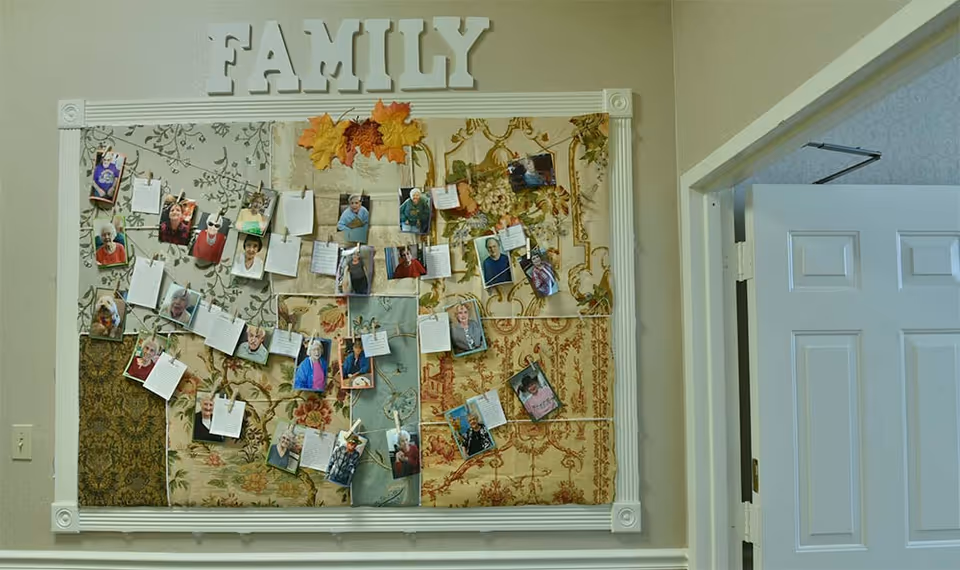 A decorative bulletin board with a patchwork fabric background displaying multiple photographs and handwritten notes clipped to strings. The board is framed in white and has the word 'FAMILY' in large white letters at the top, with some autumn leaves as decoration. The board is mounted on a beige wall next to a white doorframe.