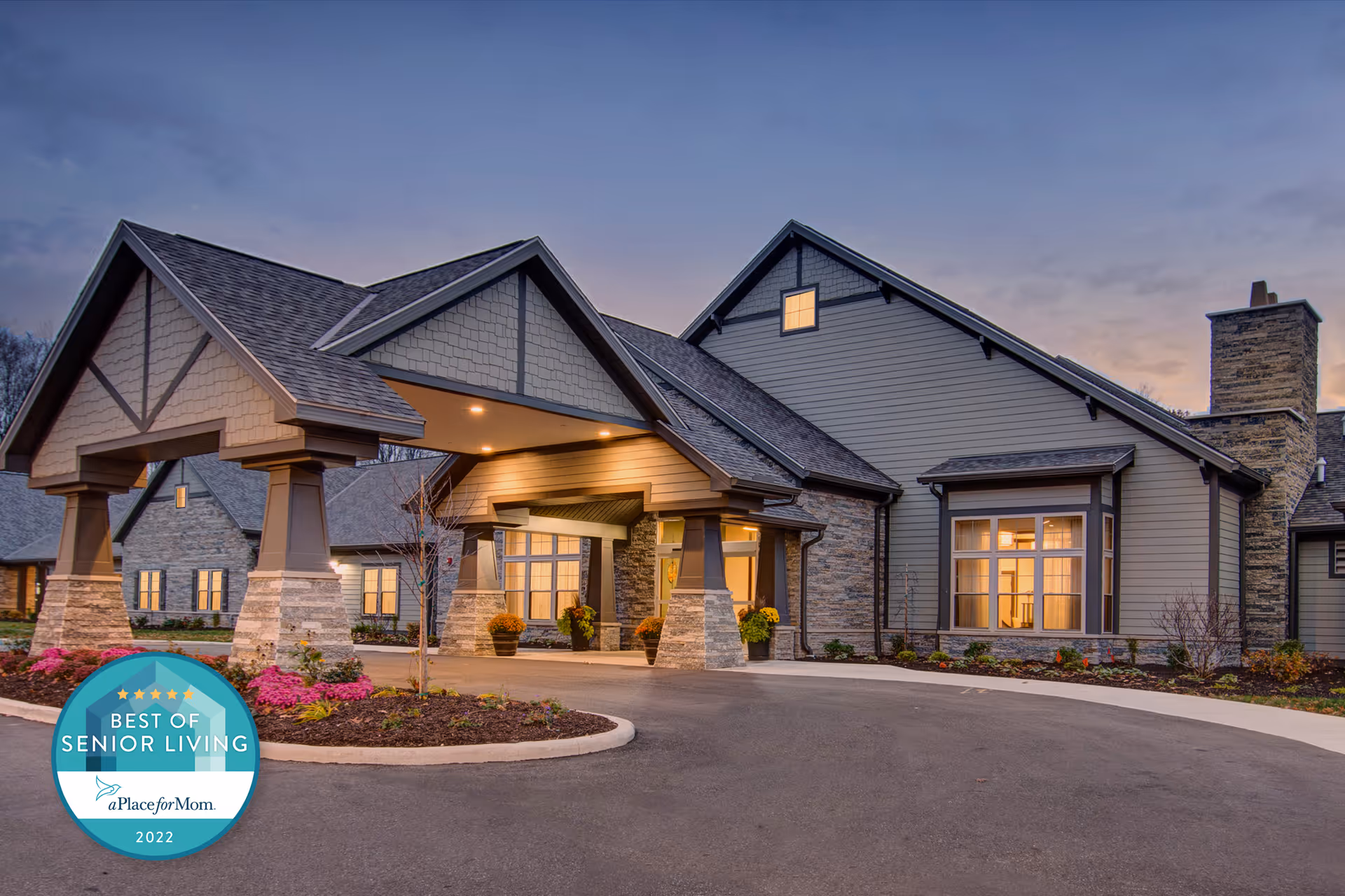 Exterior front entrance of a senior living building with a covered porte-cochere, stone columns, lit windows, and landscaped beds at dusk.