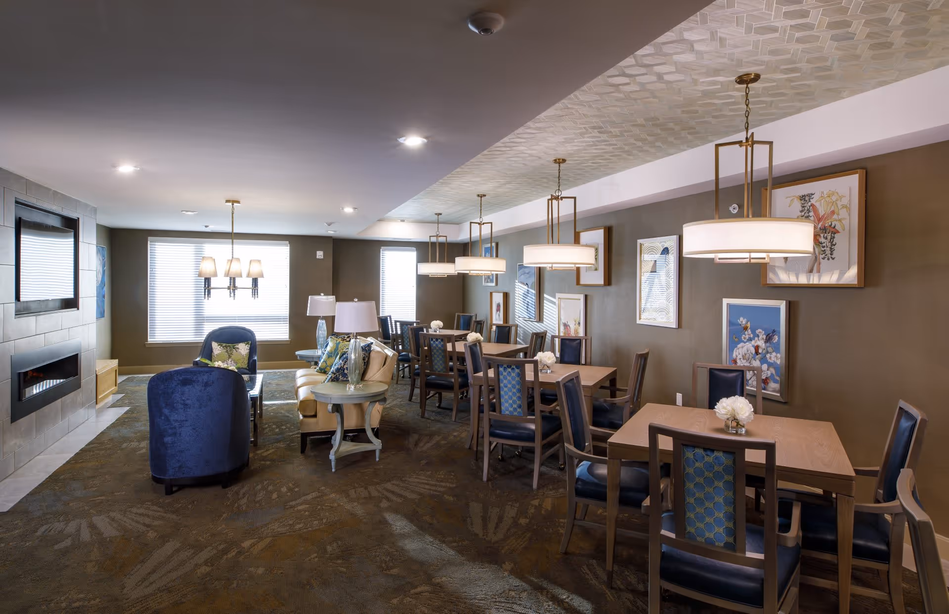 A spacious and well-lit common area in an assisted living facility featuring multiple wooden tables with chairs, decorative pendant lights hanging from the ceiling, framed artwork on the walls, a seating area with blue upholstered chairs and a beige sofa, and a modern fireplace on the left side.