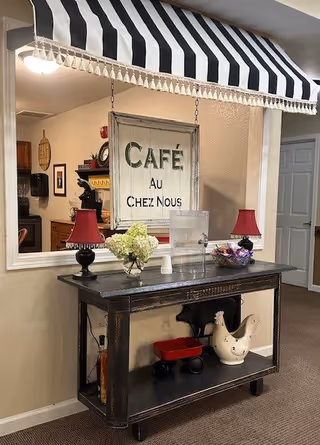 Interior view of a cozy café-themed corner in a senior living facility with a black and white striped awning above a dark wooden console table. The table holds two red lamps with black bases, a vase with white flowers, a clear container, and a bowl with wrapped candies. Below the table are decorative items including a small red wagon and a white ceramic rooster. A framed sign on the wall reads 'CAFÉ AU CHEZ NOUS'.