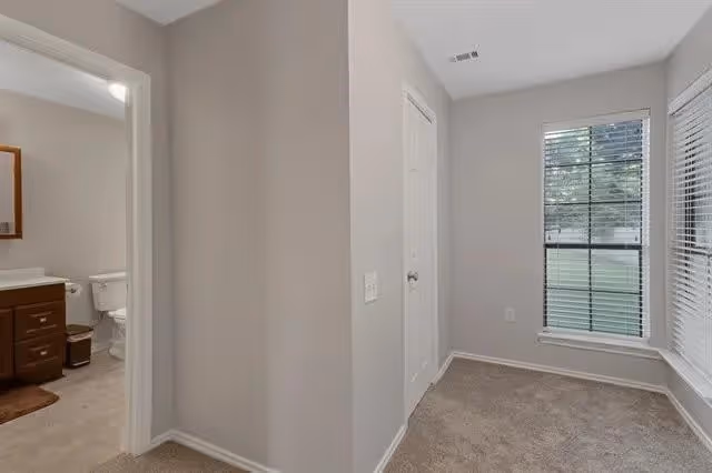 View of a small carpeted room with light gray walls and two windows with white blinds. To the left, there is an open doorway leading to a bathroom with a toilet, a wooden vanity with a sink, and a mirror above it.