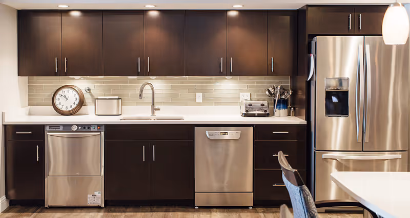 Modern kitchen with dark brown cabinets, stainless steel dishwasher, refrigerator, and a countertop with a sink, clock, toaster, and utensil holder.