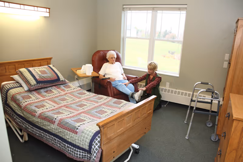 An elderly woman sitting in a red recliner chair in a bedroom, with a caregiver kneeling in front of her, helping with her footwear. The room has a wooden bed with a patterned quilt, a walker, a wooden dresser, and a window showing a green outdoor view.