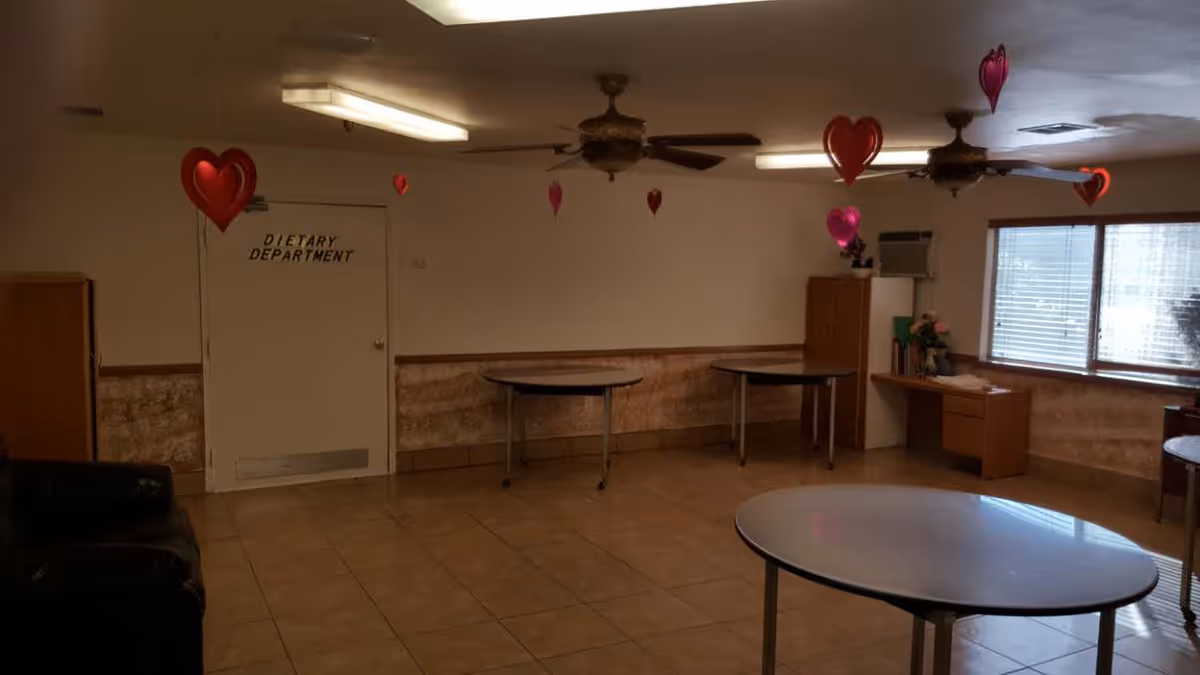 A room with tiled floor and beige walls, decorated with red and pink heart-shaped balloons hanging from the ceiling. There are three round tables, a wooden desk with flowers and books near a window with blinds, and a door labeled 'Dietary Department'. Ceiling fans and fluorescent lights are mounted on the ceiling.