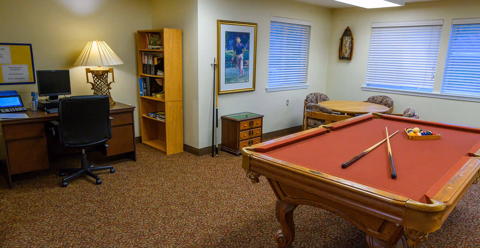 A recreational room featuring a red felt pool table with two pool cues and a rack of balls on it. To the left, there is a wooden desk with a computer, a lamp, and a black office chair. Next to the desk is a wooden bookshelf with books and games. In the background, there is a small round table with four cushioned chairs near two windows with blinds. The walls are light-colored and decorated with a framed picture and a wall clock.