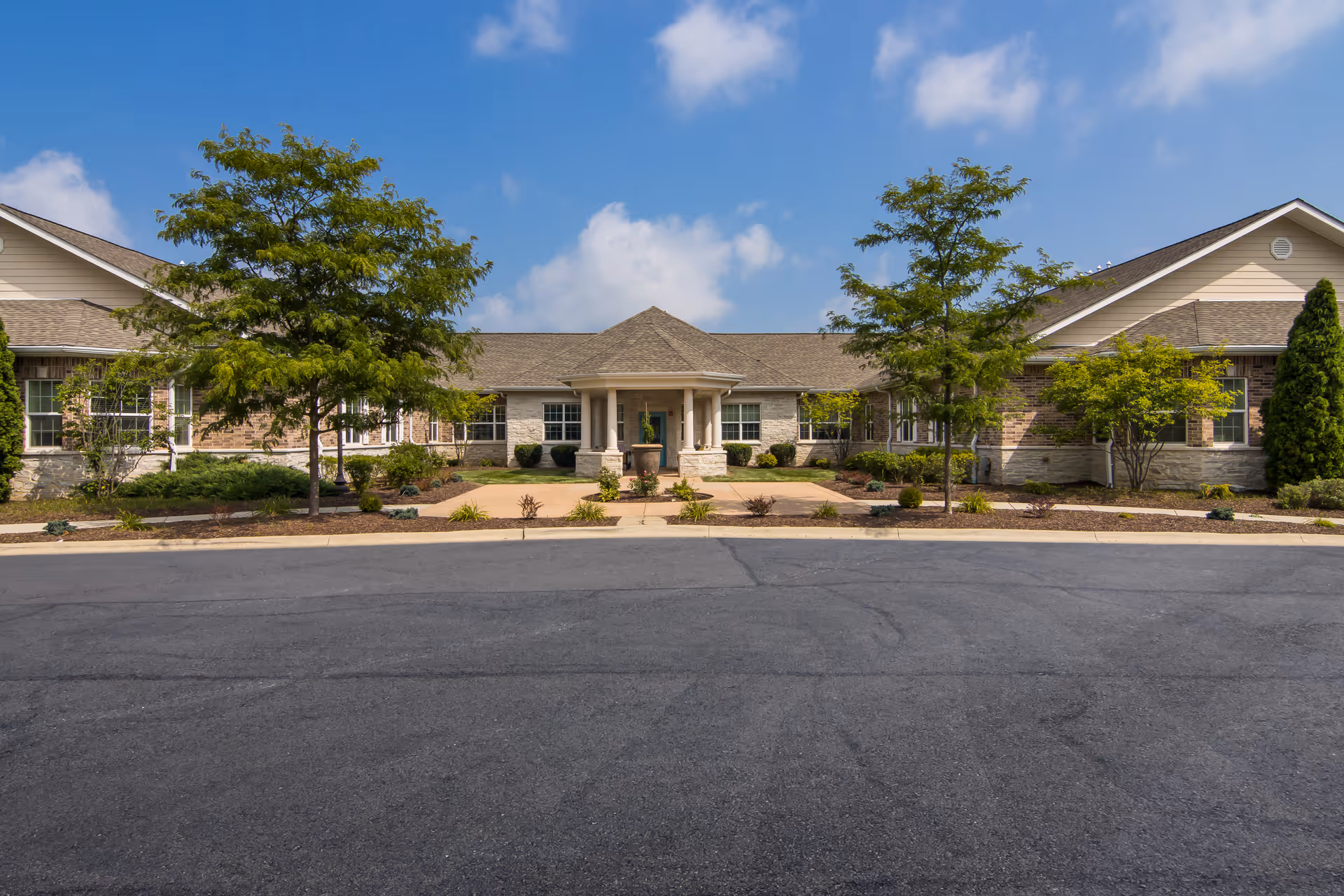 Single-story brick and siding memory care building with a central covered entrance, landscaped beds and trees under a blue sky.