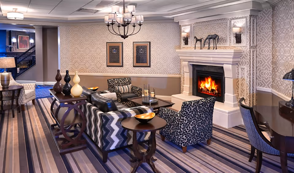 A cozy living room area in an assisted living facility featuring a lit fireplace with decorative sculptures on the mantel, patterned wallpaper, a chandelier, and comfortable seating including a chevron-patterned sofa and two patterned armchairs. The room has a striped carpet and various tables with decorative items, creating a warm and inviting atmosphere.