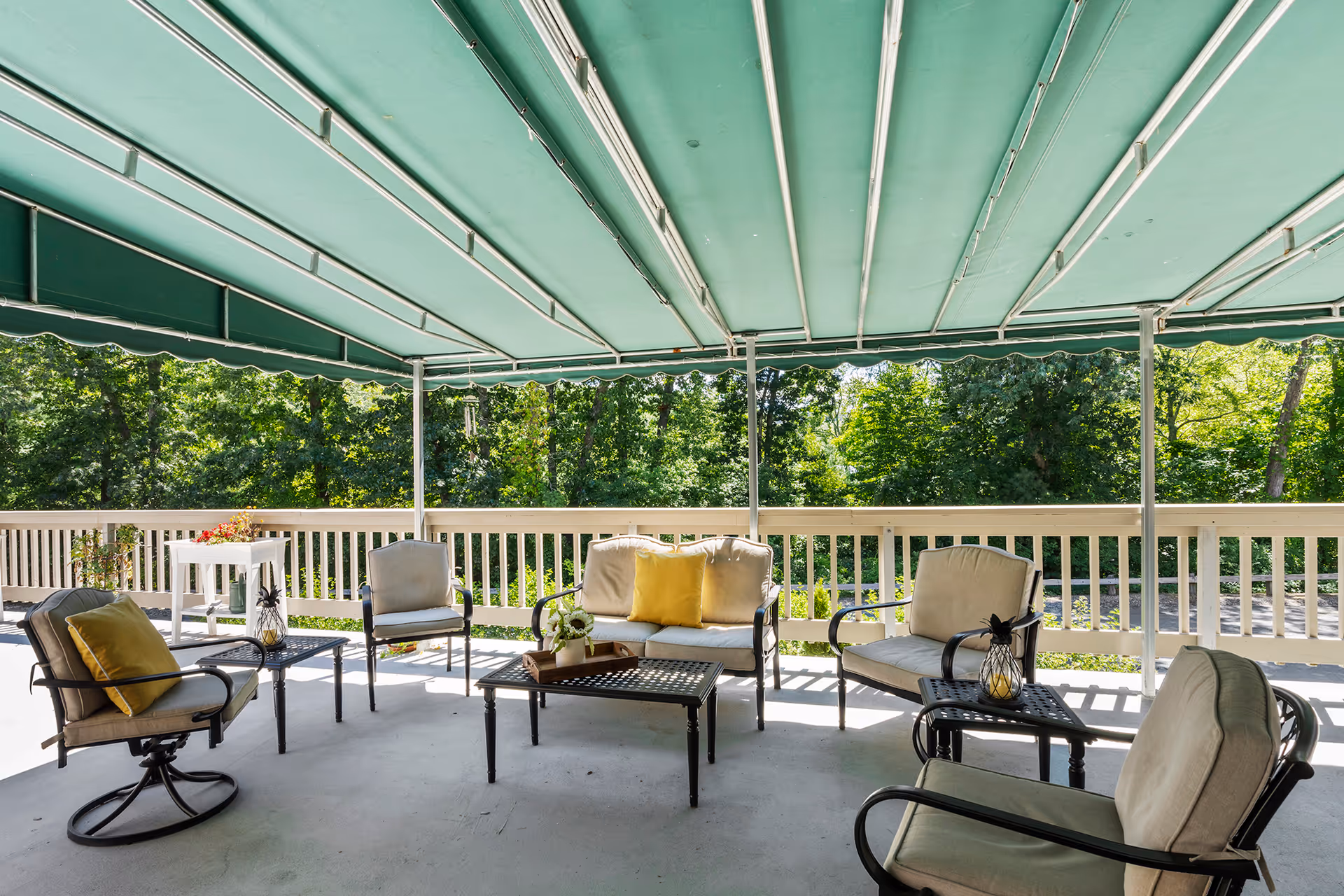 Outdoor covered patio area with cushioned chairs and sofas arranged around coffee tables, green retractable awnings overhead, and a view of lush green trees beyond a white railing.