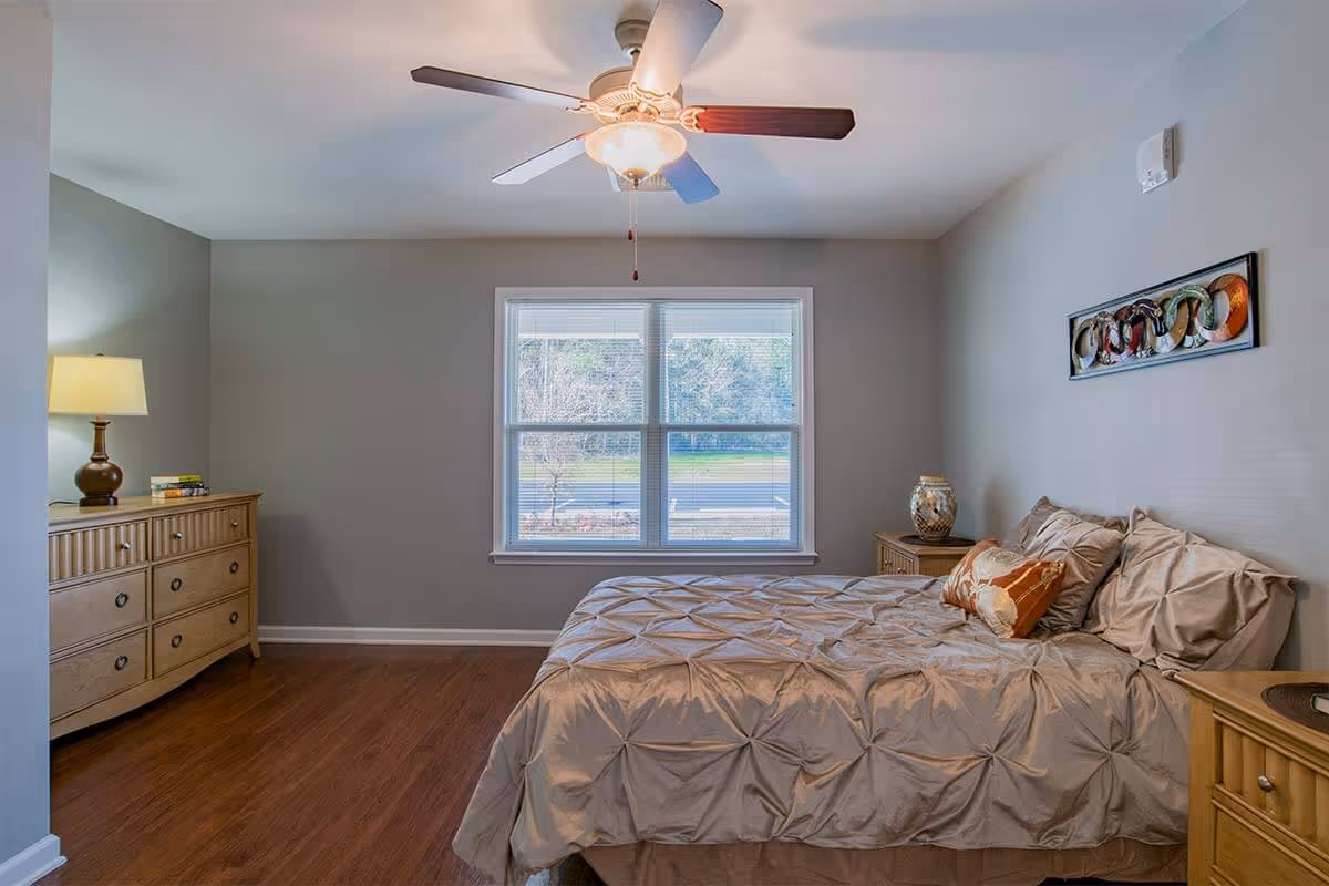 A bedroom with a large bed covered in beige bedding and pillows, two wooden nightstands on either side of the bed, a wooden dresser with a lamp and books on it, a ceiling fan with a light, and a window showing an outdoor view.