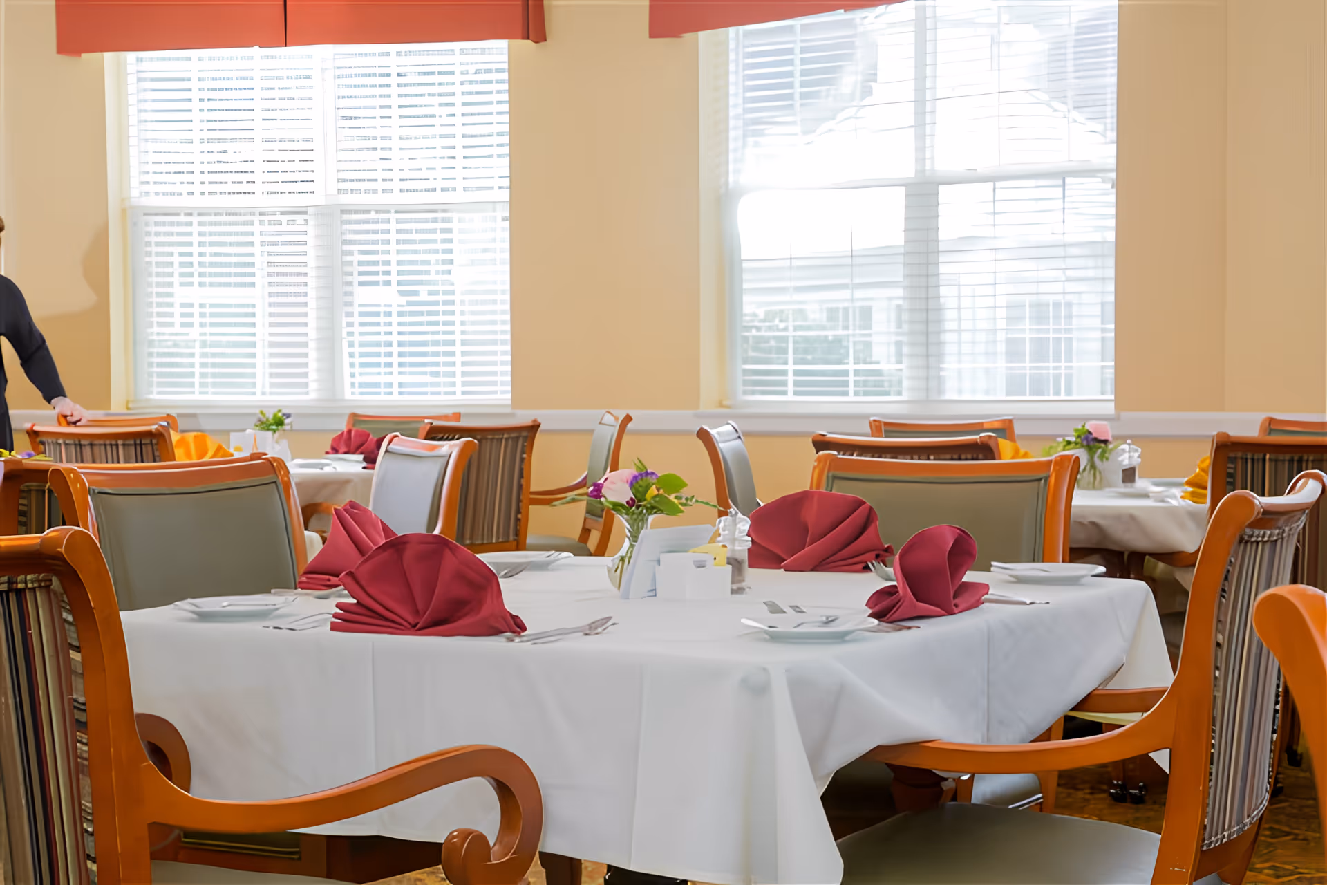 A bright dining room with multiple tables covered in white tablecloths, each set with plates, silverware, and folded burgundy napkins. Wooden chairs with cushioned seats surround the tables. Large windows with white blinds and red valances allow natural light to fill the room.