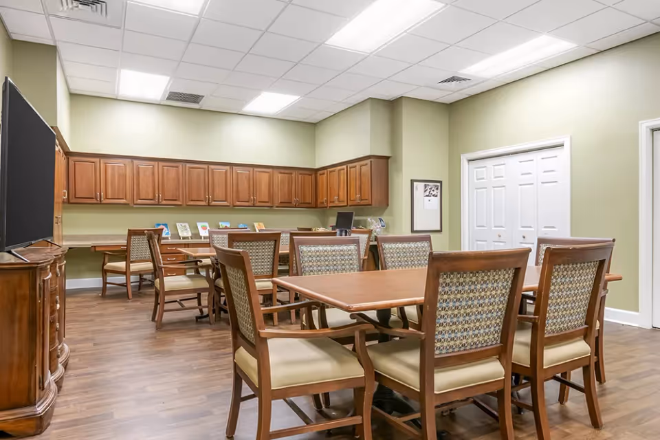 A well-lit room with wooden tables and cushioned chairs arranged for group seating. The walls are painted light green, and there are wooden cabinets mounted on the far wall. A large flat-screen TV is positioned on a wooden stand to the left. The floor is wood laminate, and the ceiling has recessed lighting panels.