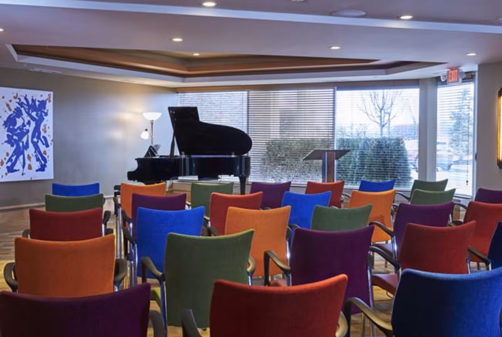 A small auditorium or meeting room with colorful chairs arranged in rows facing a black grand piano and a podium. Large windows with blinds allow natural light to enter the room, and a modern abstract painting is visible on the wall to the left.