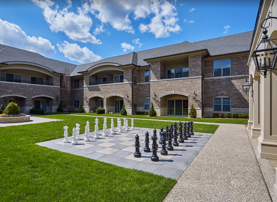 Courtyard with a large outdoor chessboard on tiled squares in front of a two-story brick building under a blue sky.