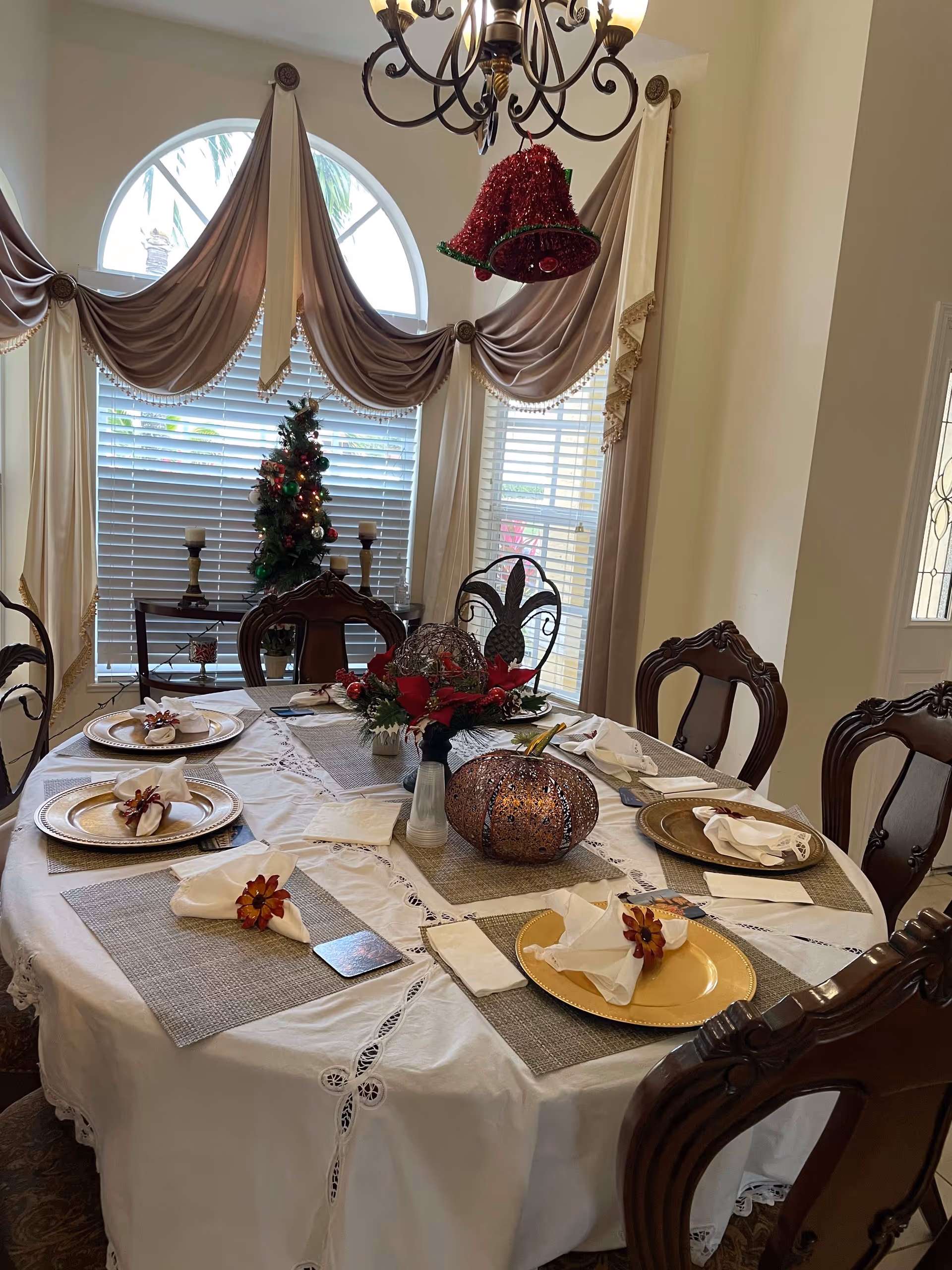 A decorated dining room with a round table set for six, holiday centerpiece and place settings, a chandelier overhead and a small Christmas tree by a curtained window.