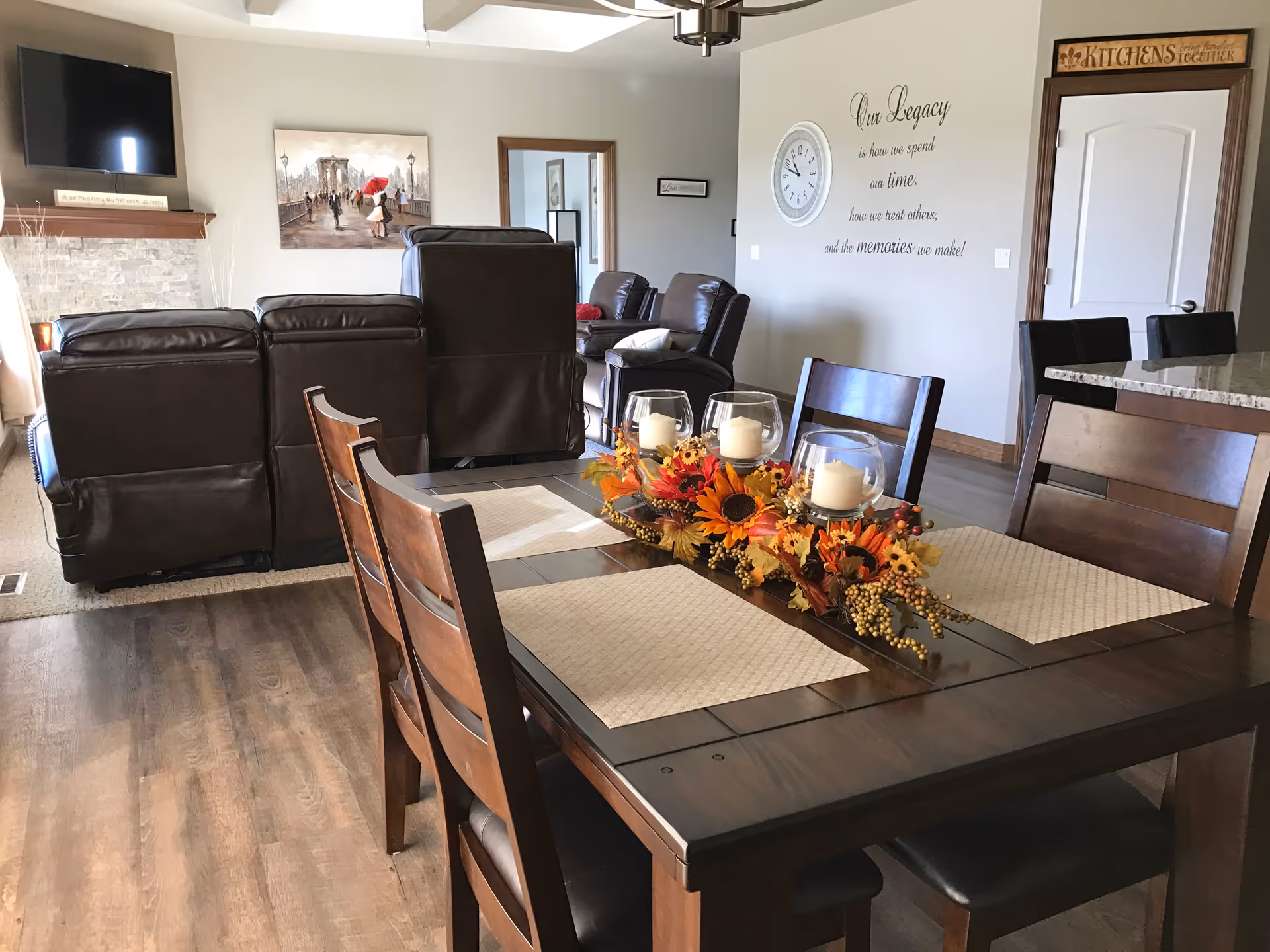Interior view of a senior living facility showing a dining table with four wooden chairs and a centerpiece of candles and autumn flowers. In the background, there are brown leather recliners facing a wall-mounted TV above a stone fireplace. A wall clock and a decorative wall quote about legacy are visible on the wall, along with a door and a kitchen counter with bar stools.