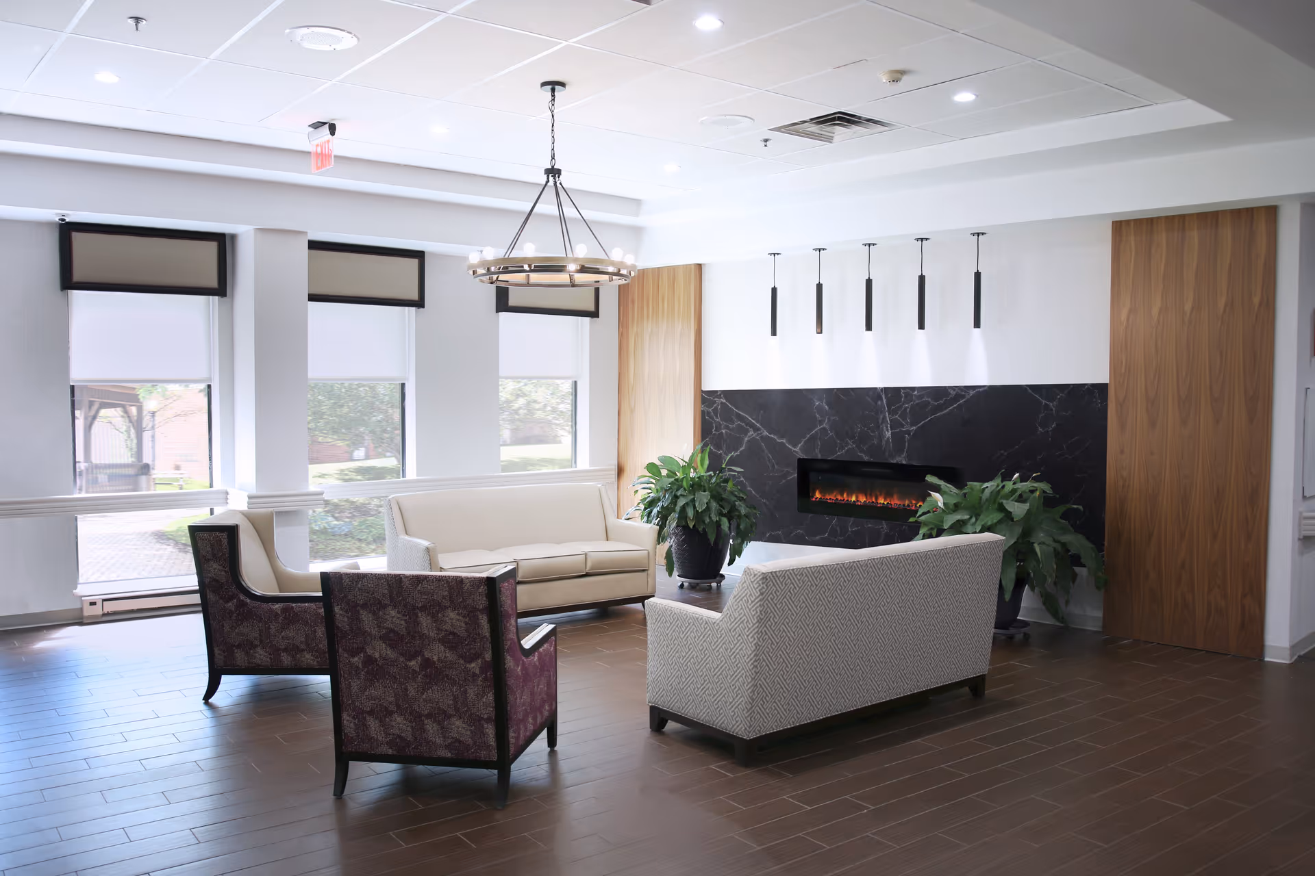 Bright communal seating area with sofas and armchairs arranged around a modern wall-mounted fireplace and potted plants.