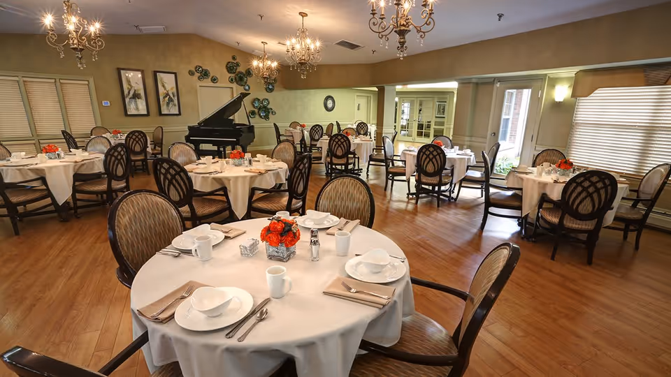 Bright dining room with round tables set for meals, upholstered chairs, chandeliers and a grand piano.